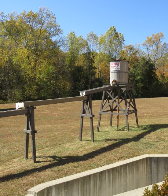 An eight-foot water tower with a wooden aqueduct leading downhill toward an activity area where kids can pan for gems and fossils under supervision.