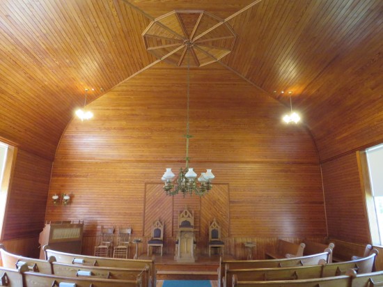A church auditorium, wood paneling covering EVERYTHING. A chandelier hangs on a long chain from a vaulted ceiling; three throne-like chairs and a podium are on the small stage.