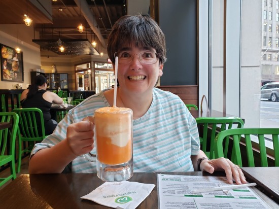 Anne with a huge smile hoisting a mug of orange Creamsicle. On the table is a Wahlburgers menu.