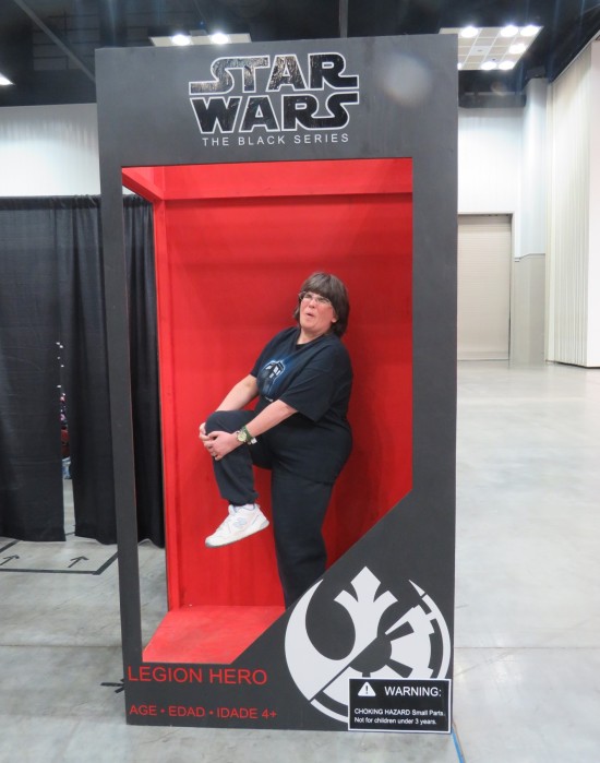 Anne standing inside a giant-sized "Star Wars: The Black Series" action figure box, holding her right foot and grimacing in pain. The bottom of the box calls her figure a "Legion Hero".
