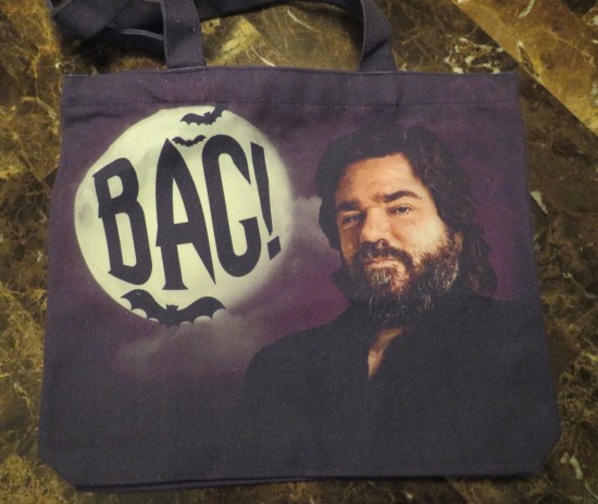 A purple and black tote bag with the painted face of Matt Berry as his character Laszlo from "What We Do in the Shadows". Next to his head, the word "BAG!" is written on a large, full moon.