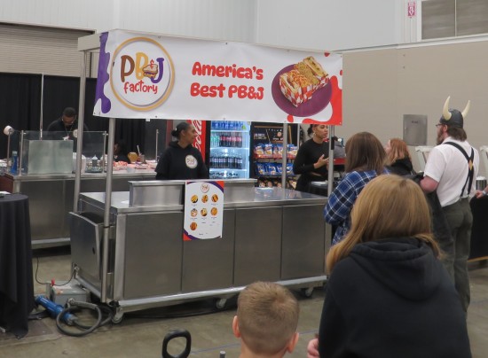 The PB&J Factory metal food-service booth with signage in whimsical font. At far right, the next customer in line wears suspenders and a Viking helmet.