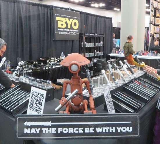 A booth selling DIY lightsabers with a Pit Droid statue on one corner, holding a lightsaber. Sign in front of droid reads "May the Force Be With You".