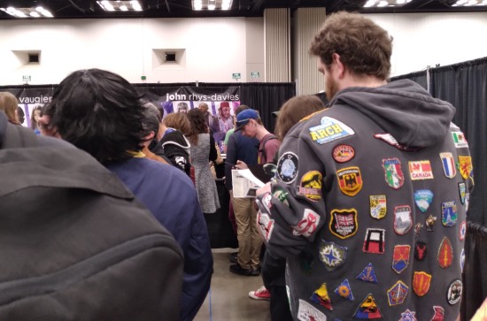 Fans lined up at the booth of John Rhys-Davies before he arrived. His banner has pics of Gimli and Sallah. The fan in front of us wears a hoodie covered in travel and geek patches.