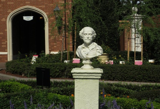 A Shakespeare bust on a 5-foot-tall pedestal standing in the middle of a garden and manicured bushes along walkways. Flowers are purple and yellow-orange.