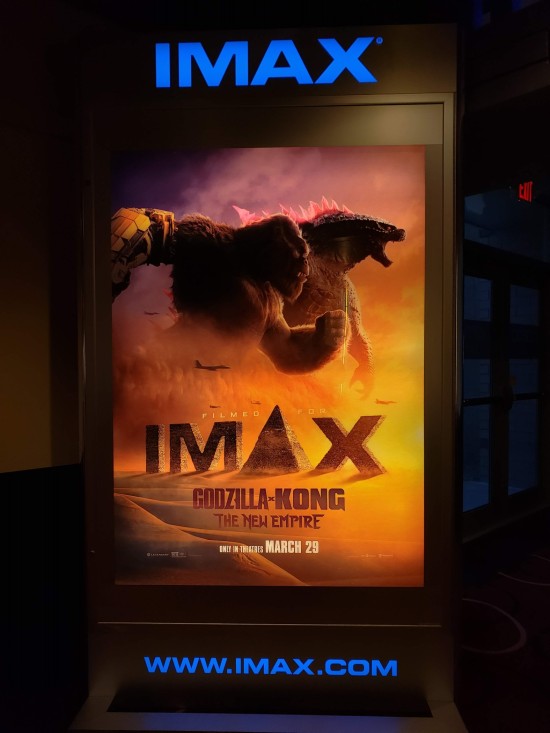 The yellow-and-black IMAX movie poster for "Godzilla x Kong: The New Empire". The title monsters are running in shadowed profile. Tiny fight jets zoom alongside them. The 'A' in "IMAX" is replaced with a Pyramid thinner than any real Egyptian Pyramid.