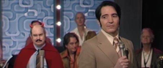 1970s TV show host holds a mic and side-eyes stage right. Behind him is his house band, led by a chubby bald guy wearing red devil horns and a cape for Halloween.