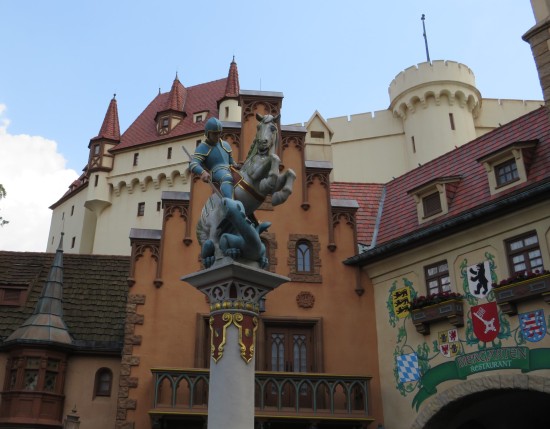 Statue of a blue knight and a gray horse atop a tall, narrow pedestal in the center of a life-size replica German town square. 