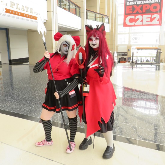 Two women in red, black and white costumes with staves. C2E2 sign is in the upper-right corner.